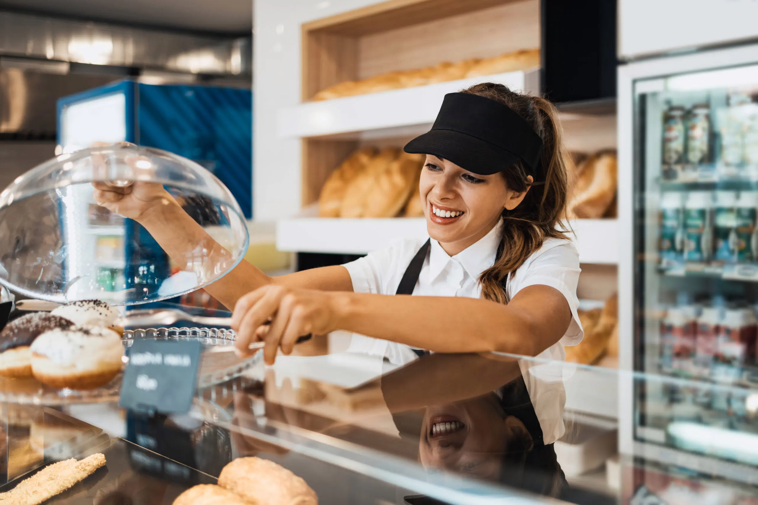 Convenience store staff member reaches for fresh pastries beside refrigerated food and drink displays.