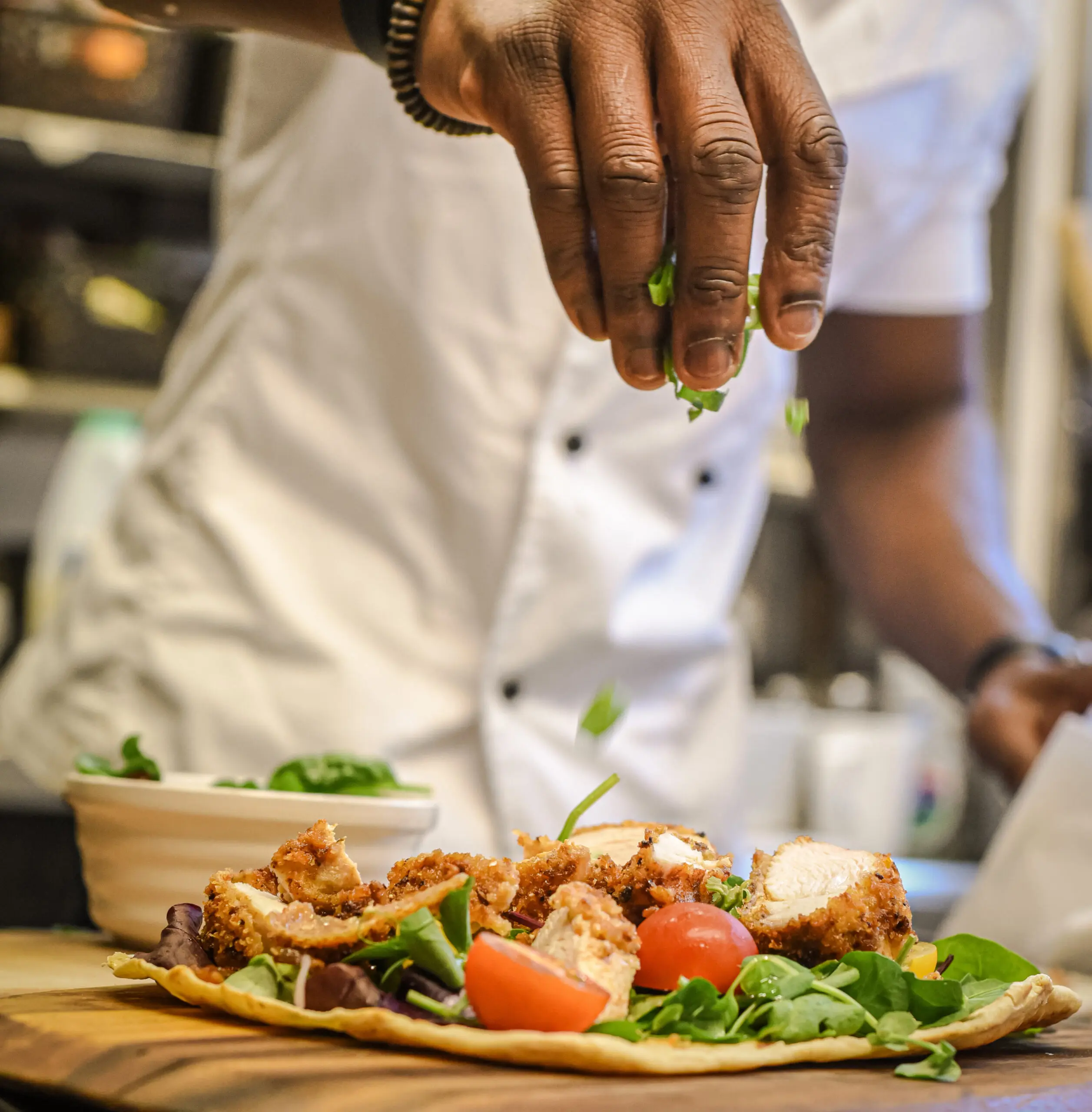 Chef sprinkles fresh green herbs over a crispy chicken flatbread topped with greens and tomatoes.