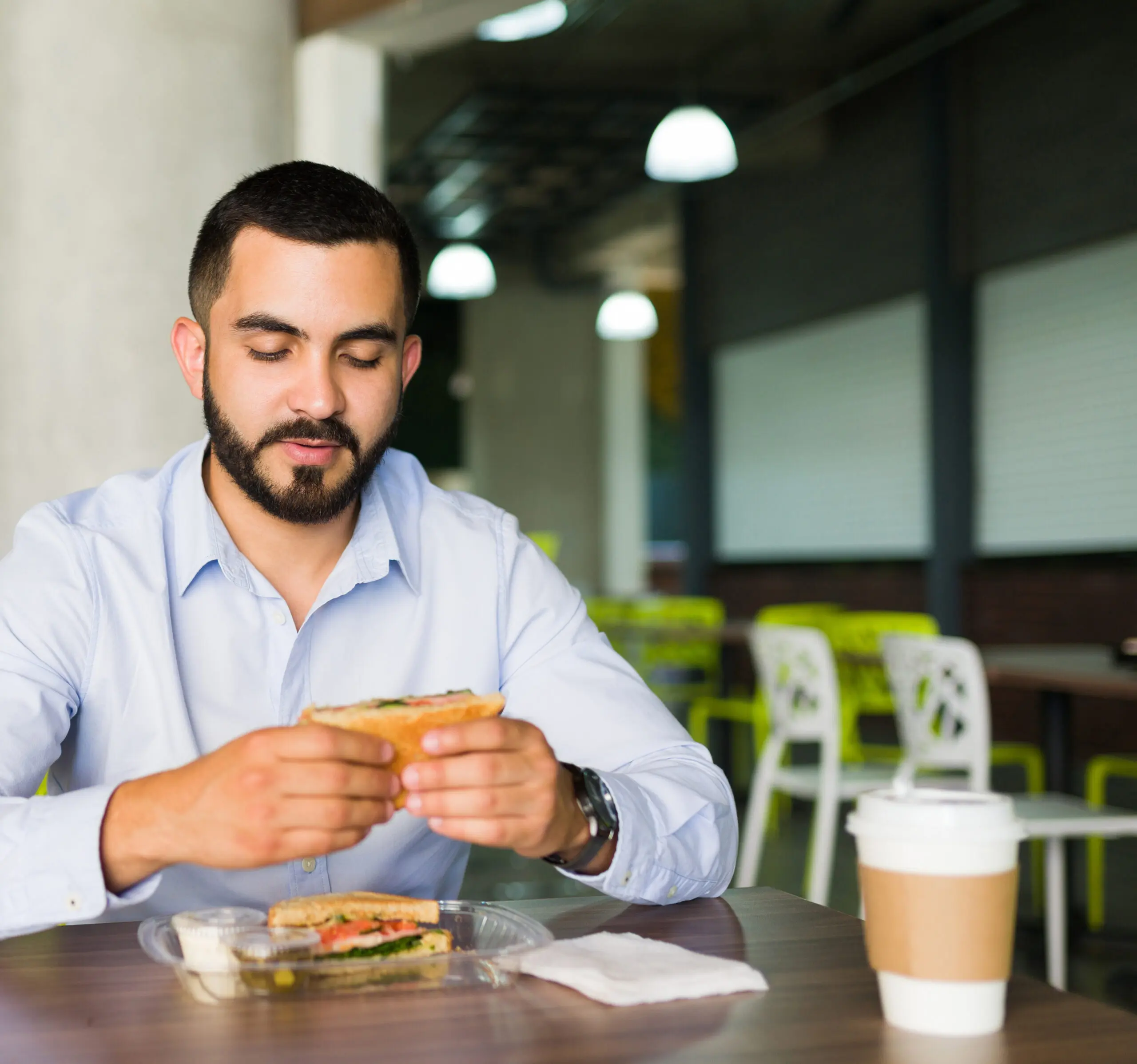 Man seated at a table in a cafeteria dining space, holding a sandwich.